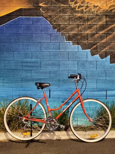 A rebuilt orange Malvern Star step through bicycle standing against a painted blue brick wall